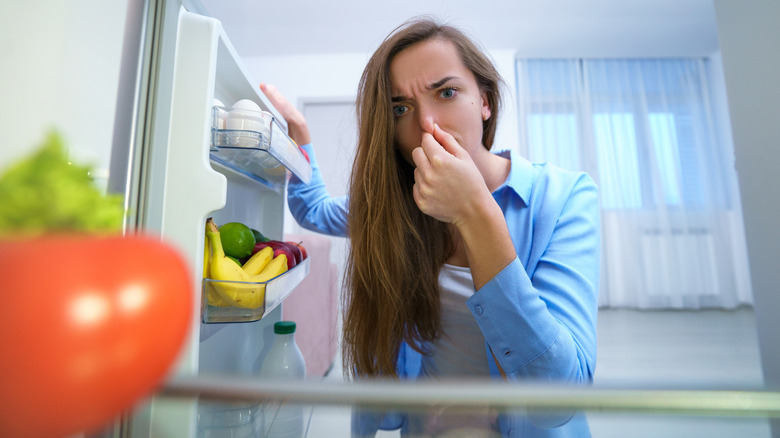 женщина закрывает нос рукой от неприятного запаха, исходящего из открытого холодильника